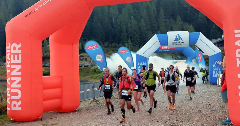 Een groep hardlopers in startnummers begint aan een trailrun en loopt onder een grote rode opblaasbare boog door, met blauwe spandoeken en een beboste berg op de achtergrond.